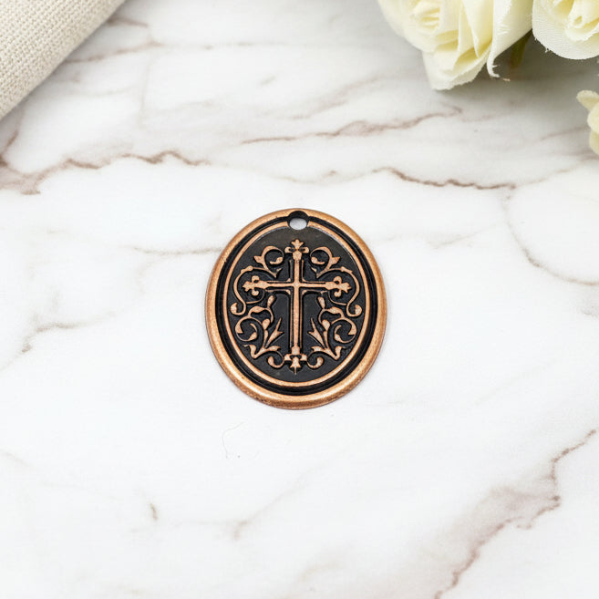 A bronze cross pendant with intricate designs on the arms, placed on a white surface with a blurred background of yellow flowers.