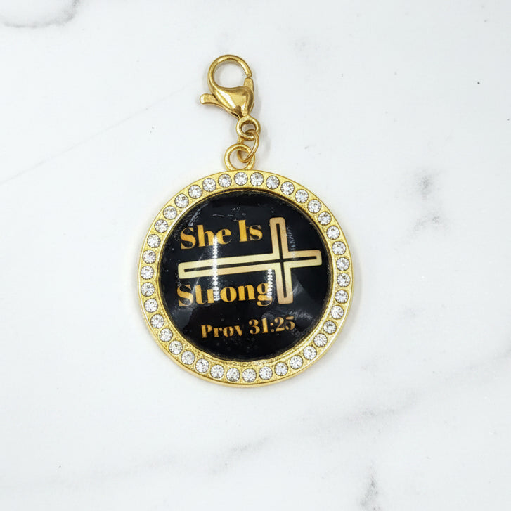 A gold-colored charm with a black background featuring a cross and the text 'SHE IS' along with a reference to 'Proverbs 31:25'.