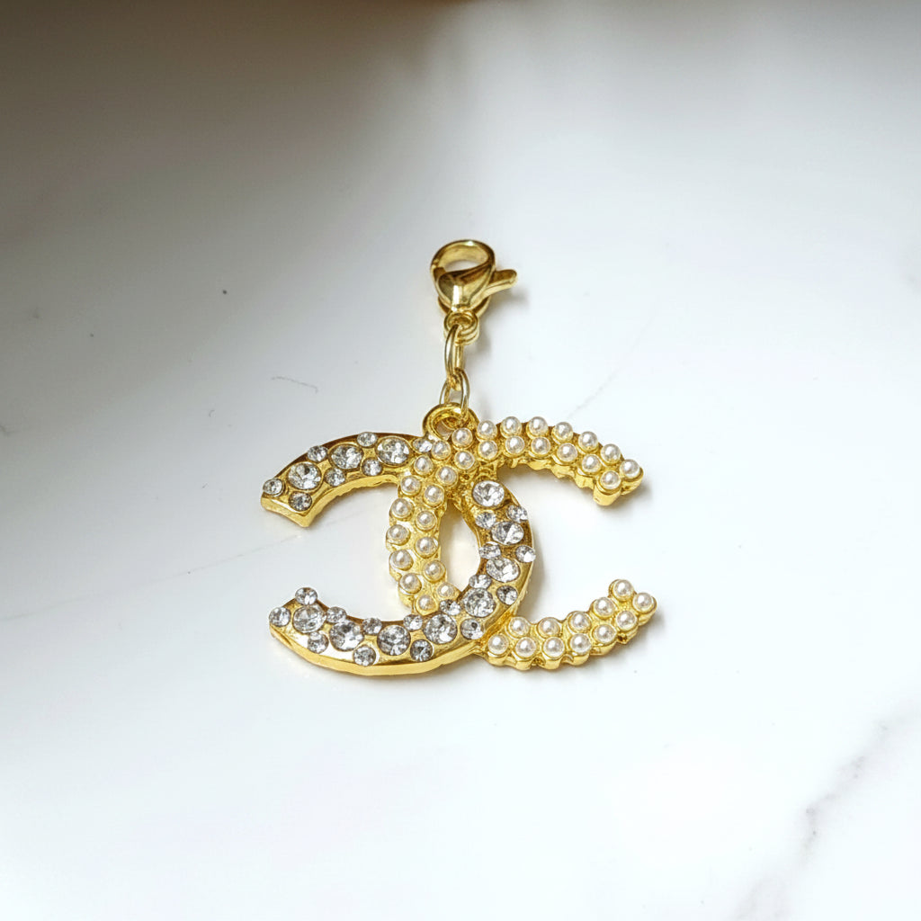 A Chanel CC inspired charm with a gold-colored metal base and a chain of pearls, surrounded by clear, round diamonds.