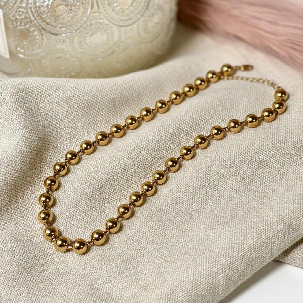 Gold beaded necklace on a linen surface with an ornate silver container in the background.