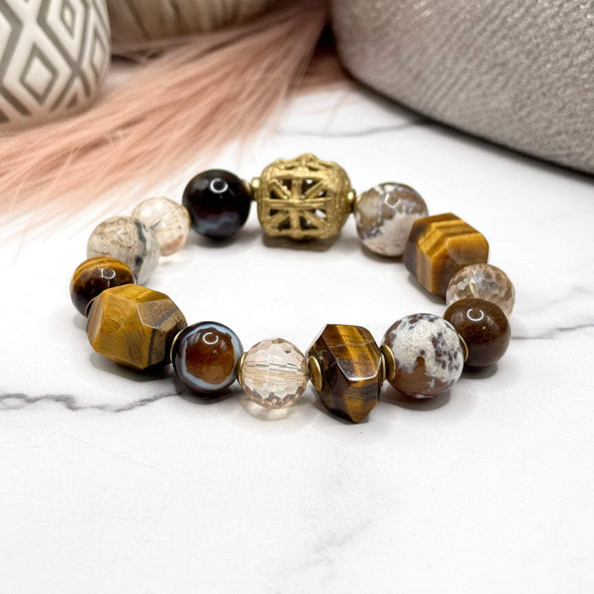 A bracelet with a mix of brown, white, and black beads, alongside a gold-colored geometric bead, displayed on a white surface next to a gray textured object.
