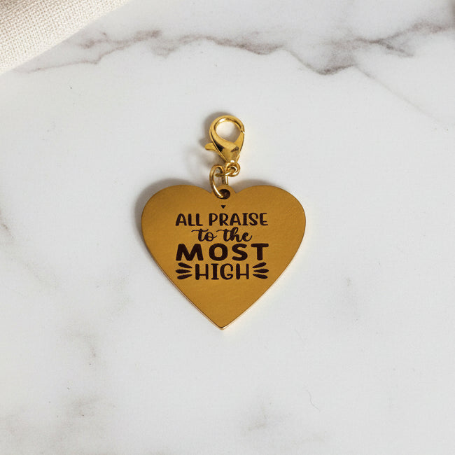 A gold-colored metal heart-shaped charm with the inscription 'All Praises'.