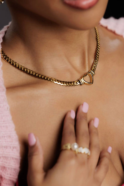 Gold chain necklace with a heart clasp worn by a person, with a hand touching the chest.