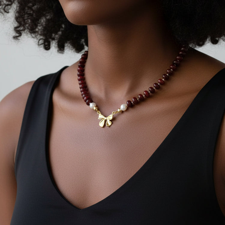 Necklace with red beads and a gold bow charm worn by a person in a black tank
