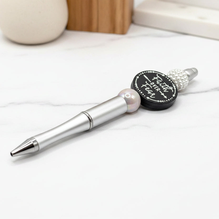 Silver pen with decorative cap featuring 'Faith Over Fear' text, next to a ball of yarn on a light background.